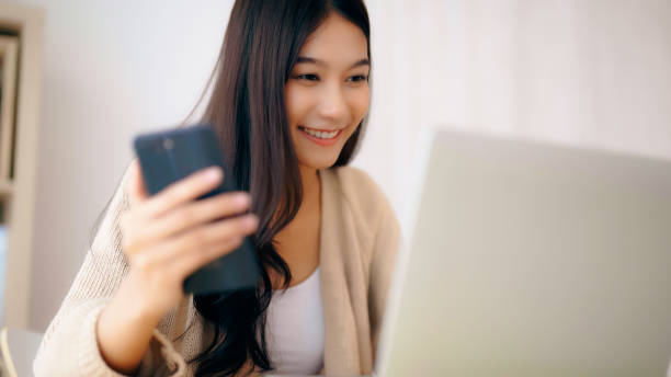 istock Young asian woman happy working on computer laptop and hand typing chat message on mobile smartphone at home. Video call, Online meeting 1474940927