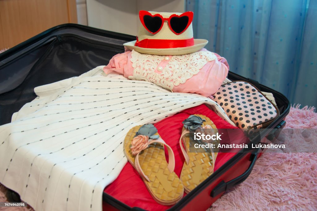Preparation for summer holidays vacations: open red suitcase with floral slippers shoes, female beach clothes, straw hat, cosmetic bag and sunglasses on bed. Packing things for journey traveling. Preparation for summer holidays vacations: open red suitcase with floral slippers shoes, female beach clothes, straw hat, cosmetic bag and sunglasses on bed.

Packing things for journey, traveling, trip or voyage. Adventure Stock Photo