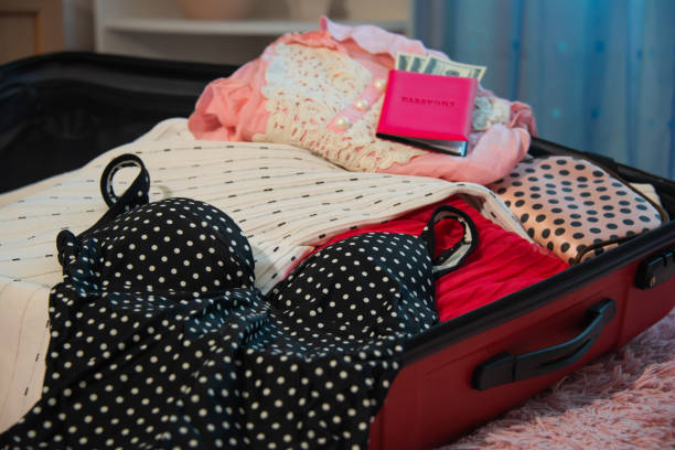 Preparation for summer holidays and packing suitcase for traveling trip: open red case bag with black swimsuit with polka dots, female beach clothes, pink passport with dollars money and cosmetic bag.