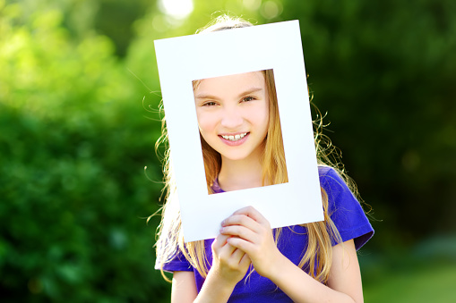 Cute cheerful little girl holding white picture frame in front of her face. Adorable child framing her smiling face.