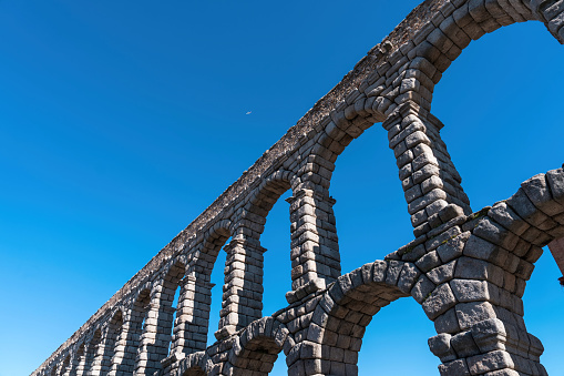 Skyward view of the beautiful arches of the Roman aqueduct of Segovia Spain and a tiny white airplane against a bright blue summer sky