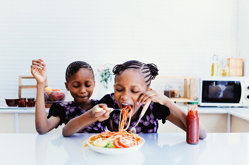 Twin girls enjoy eating spaghetti in kitchen