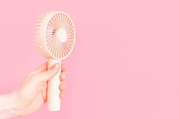 Photo of Female hand holding a small fan on pink background close-up.