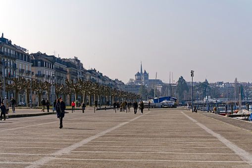 Tourists walking at quay named Gustave Ador with tree alley at lake shore of Lake Geneva on a sunny late winter day. Photo taken March 5th, 2023, Geneva, Switzerland.