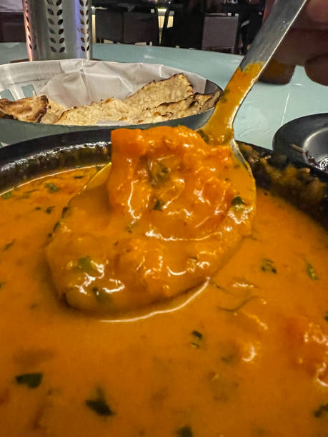 Close-up image of restaurant table setting of metal spoon over black cast iron pan containing prawn vindaloo curry, background of bowl of naan (flatbreads), focus on foreground stock photo