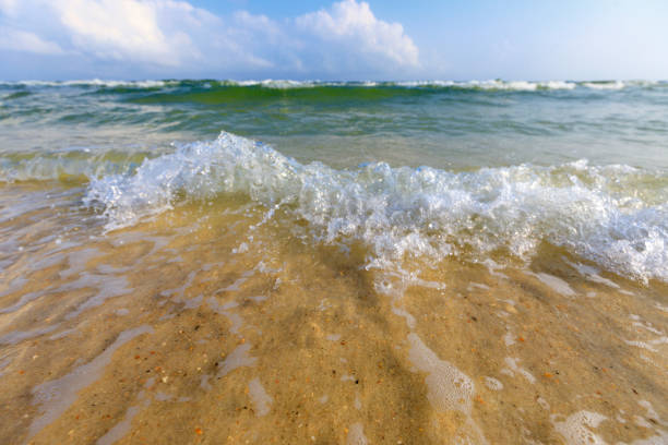 low macro view of waves crashing down on the beach with clear green water, light tan sand, blue sky and a cloud.
