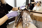 Back view of woman sitting in front of mirror in beauty salon. Hairdresser colorist taking off foil from strand of hair.