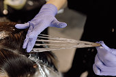 Unrecognizable woman hairdresser colorist hold strand of long hair, distributing hair dye with fingers in blue gloves.