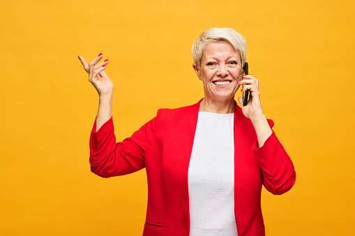 Cheerfu aged woman with short blond hair talking on mobile phone and looking at camera while standing against yellow background