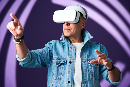 Senior man in casualwear and vr headset pointing at something while traveling in futuristic reality against lavender background