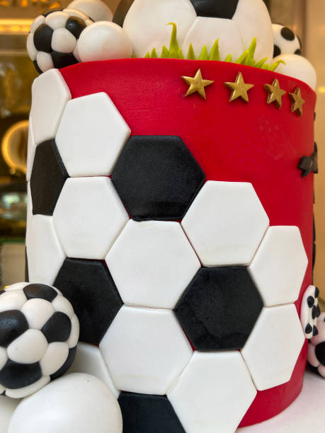 Stock photo showing close-up view of a layered birthday cake decorated with soccer theme with fondant icing in shades of red, black, white and gold. imagen en primer plano de pastel de cumpleaños en capas con temática de fútbol decorado en un tablero de pastel con azulejos teselados de glaseado de fondant de forma hexagonal en blanco y negro con fondo rojo, balones de fútbol y hojas de glaseado de