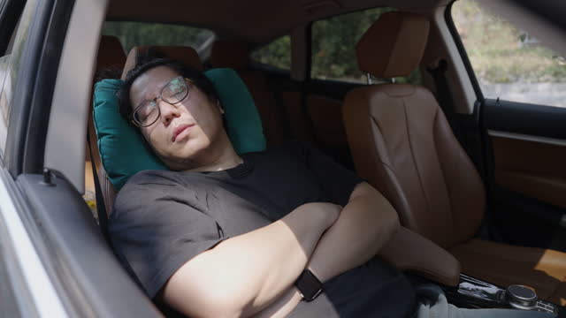 Asian large build man falling asleep while waiting for friend in a car so he's grabbing a pillow and napping with arms crossed on driver's seat.