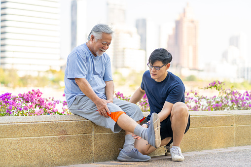 Elderly man suffering knee pain during jogging exercise with his son at park in the city. Adult man gives first aid to father with knee injury. Family relationship and older people health care concept