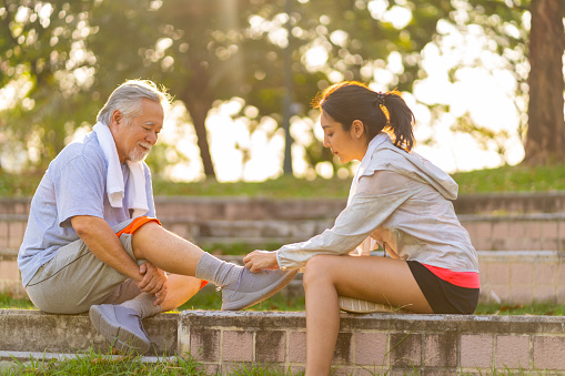 Asian woman daughter helping elderly father tie running shoe laces during jogging exercise together at park. Retired senior man health care with outdoor lifestyle sport training workout in the city.
