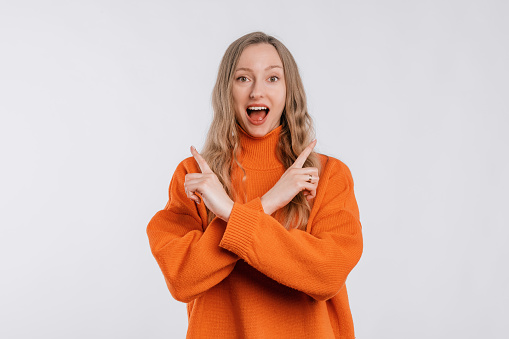 Positive friendly young woman with a happy smile on face pointing fingers sideways, showing left and right promo, stands in an soft orange sweater over neutral background. Great idea cool offer