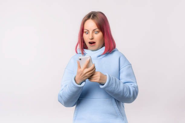 istock Surprised young woman looking at mobile phone, reading unexpected message with open mouth, checking social networks. Lovely female model with pink hair in a blue hoodie standing over white background 1474984780