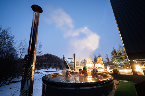 Family enjoying bathing in wooden barrel hot tub in the terrace of the cottage. Scandinavian bathtub with a fireplace to burn wood and heat water.