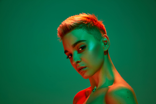 Portrait of young blonde girl with short hair and bare shoulders posing against green studio background in orange neon light. Cyberpunk culture. Concept of technology, modern fashion, futurism