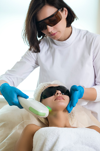 Close-up of Young Winsome Woman in UV Protective Glasses During Laser Skin care on Face Using  Rejuvenation Intense Pulsed Light Therapy IPL. Vertical image Orientation