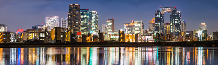 Panoramic view across the tranquil waters of the Yodo River reflecting the futuristic skyscraper cityscape of central Osaka at sunset, Japan.