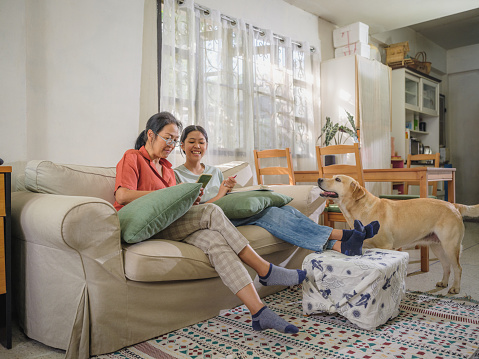 Happy Asian teenage daughter and mother using smart phone for online banking or video call while setting on sofa in living room while dog stood beside at home, Family spending time together at home, People lifestyle technology working with mobile digital devices
