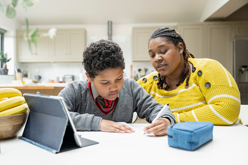 Waist-up view of 10 year old multiracial boy in school uniform sitting at kitchen counter in modern home with early 30s woman, using digital tablet and writing.