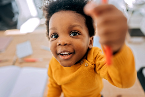 Cute little boy pointing pencil at camera while colouring book at home