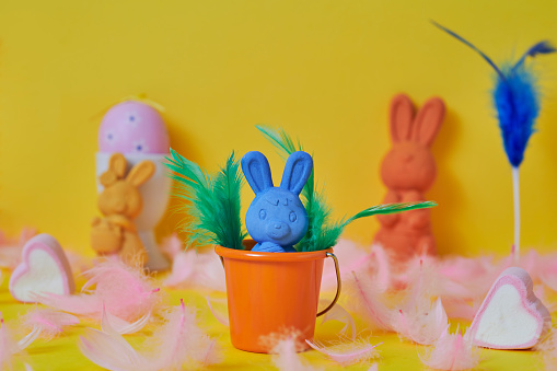a blue easter bunny in an orange bucket surrounded by pink feathers and a pink easter egg and some other easter bunnies of different colors on a yellow background