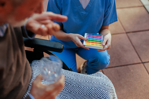 A caregiver with a pillbox is next to an elderly man taking the medication in the nursing home's yard.