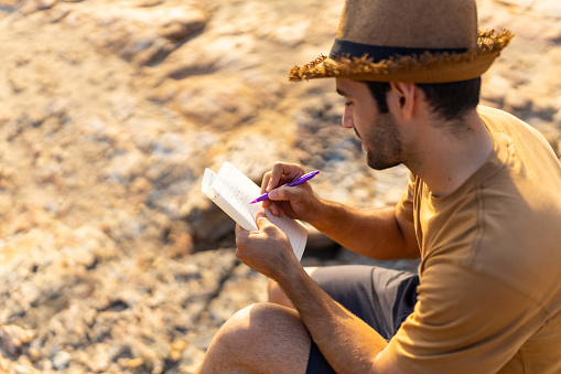 Caucasian man traveler looking to the ocean with writing journey memories in diary book on coastline hill at summer sunset. Handsome guy enjoy outdoor lifestyle hiking on island mountain on holiday vacation. Solo travel concept.