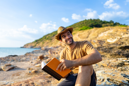 Caucasian man traveler reading a book and looking to the ocean in diary book on coastline hill at summer sunset. Handsome guy enjoy outdoor lifestyle hiking on island mountain on holiday vacation. Solo travel concept.