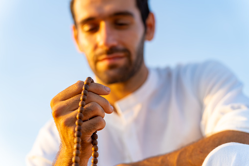 Caucasian man counting wooden mala beads practice meditation prayer with ocean on rocky coastal hill at summer sunset. Mental health care, self motivation, outdoor relaxing and nature therapy concept.