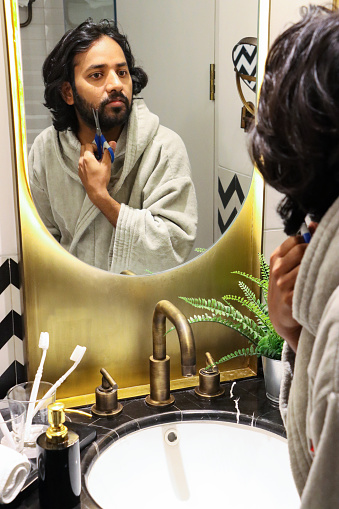 Stock photo showing luxury bathroom, with black and white zig-zag patterned wall tiles, white ceramic sink with dual knob monobloc mixer tap in front of mirror reflecting an Indian man wearing grey bathrobe whilst using scissors to trim his facial hair. Self care and grooming concept.