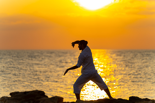 Caucasian man martial art practice and meditation with ocean nature on rocky coastal hill at summer sunset. Wellness people do outdoor relaxing yoga exercise. Health care and self motivation concept.