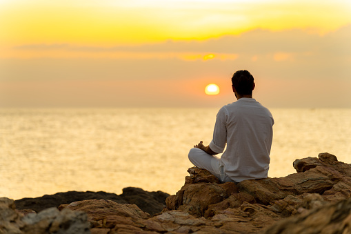 Caucasian man practicing meditation prayer and yoga exercise with ocean on coastal hill at summer sunset. Wellness health care, self motivation, outdoor lifestyle relaxing and nature therapy concept.