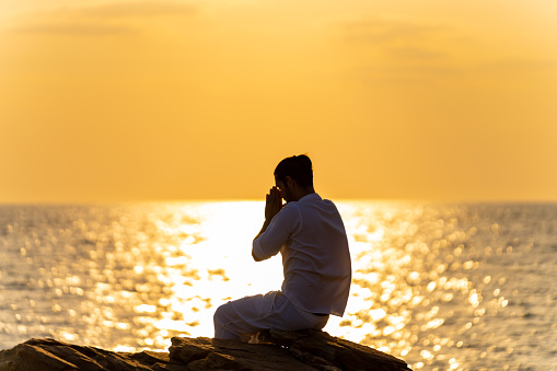 Caucasian man practicing meditation prayer and yoga exercise with ocean on coastal hill at summer sunset. Wellness health care, self motivation, outdoor lifestyle relaxing and nature therapy concept.