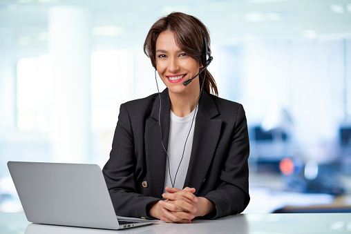 Customer service assistant wearing headset while sitting behind her computer and working in the call center. Attractive business woman wearing black blazer.