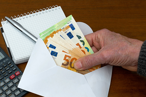 Man's hand holding european banknotes from an envelope. On the table are a calculator and a notepad with a pen.