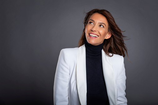 Close-up of an attractive middle aged woman with toothy smile wearing white blazer while sitting at isolated dark background. Copy space. Studio shot. Woman looking up.