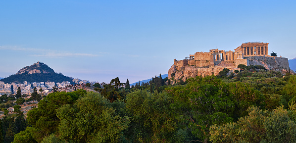 View of Acropolis hill with Propylaea and Parthenon and hill of Lycabettus in background in Athens, Greece from Pnyx hill in soft summer sunset sky. Great fresh foliage of olives and cedar trees in foreground