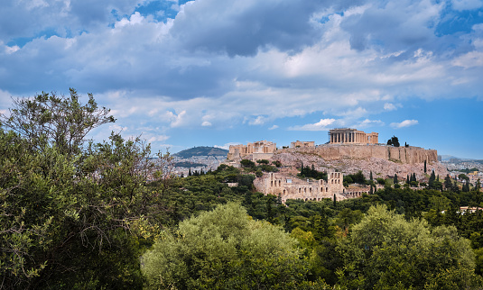 View of Acropolis hill with Propylaea and Parthenon and theater of Odeon below it in Athens, Greece from the hill of Philoppapos or Muses in summer daylight with great clouds in blue sky. Great fresh foliage of olives and cedar trees. Wide shot