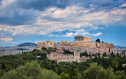 View of Acropolis hill with Propylaea and Parthenon and theater of Odeon below it in Athens, Greece from the hill of Philoppapos or Muses in summer daylight with great clouds in blue sky. Great fresh foliage of olives and cedar trees. Wide shot