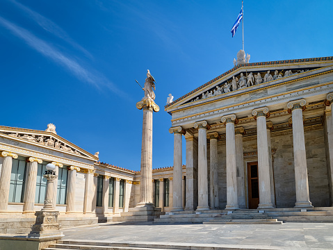 Entrance and left wing of Academy of Athens, Greece, national highest research center, and column or pillar with statue of Athena, ancient Greek goddess and patron of city, Greek flag, in bright daylight, summer sunshine. Neoclassical architecture.