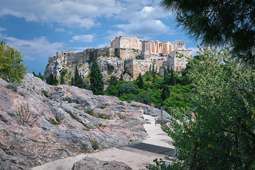 View of Acropolis hill from Areopagus hill in summer daylight with great clouds in blue sky, Athens, Greece. UNESCO world heritage. Propylaea entrance, Agrippa's monument, Parthenon. Fresh green foliage and stone and rocks of hilltop.
