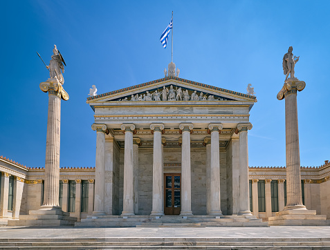 Marble main entrance of Academy of Athens, national research center, columns or pillars with Athena, ancient Greek goddess and patron of city, and Apollo, ancient Greek god and patron of arts and science. National Greek flag. Summer day, blue sky.
