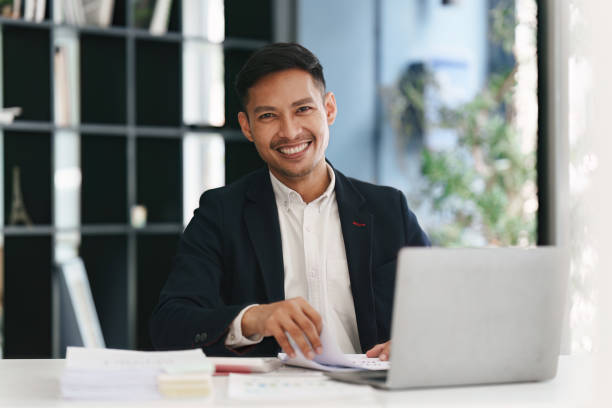 istock Smiling Asian Banker makes financial report and studies annual figures, analyzes profits. Accountant checks status of financial 1475013025