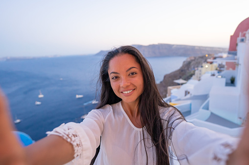 Young female asian woman with white dress on a vacation in Santorini, taking selfie on a traditional architecture background