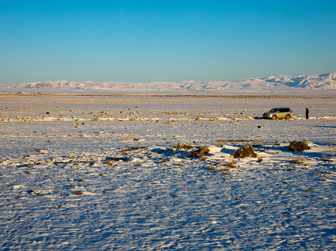 A man in a down jacket and SUV stand on the snowy alpine Chui steppe. In the distance - a mountain range, a valley. Blue sky. Travel to winter Altai mountains.