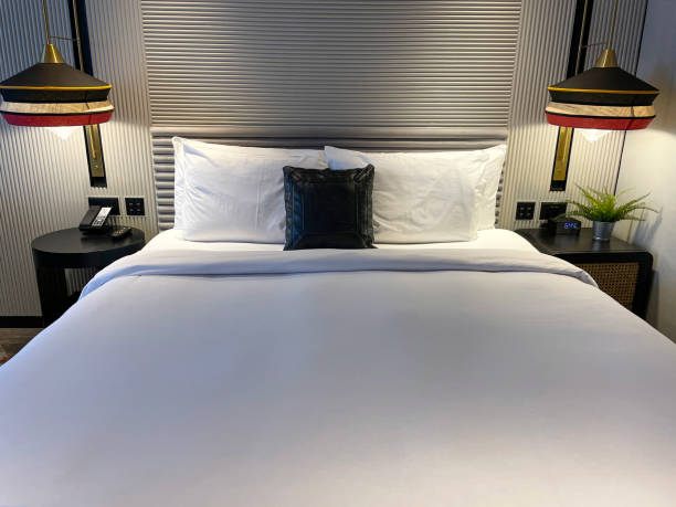 istock Close-up image of double bed with textured, baton headboard, stacked white pillows, folded duvet, digital alarm clock on nightstand with artificial fern in metal plant pot, illuminated hanging pendant lamp, beside telephone, focus on foreground 1475015061