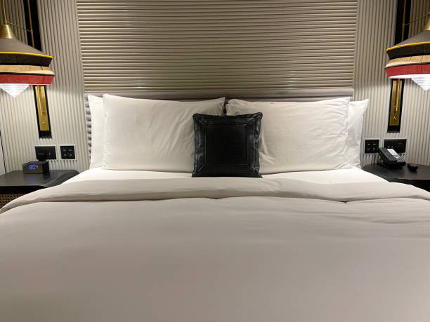 istock Close-up image of double bed with textured, baton headboard, stacked white pillows, folded duvet, digital alarm clock on nightstand, illuminated hanging pendant lamp, remote control beside telephone, focus on foreground 1475015109